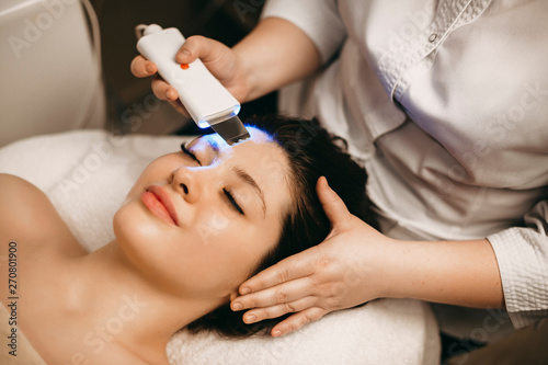 Lovely caucasian woman doing ultrasonic procedure on her face while leaning on a spa bed with closed eyes.