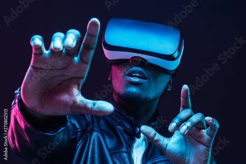 Young african man wearing virtual reality goggles with hands up, isolated on black background photo