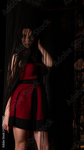 Pretty young sexy model female with dark hair in amazing long red dress and black shoes posing in dark studio