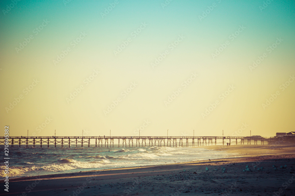 A long fishing pier heading out into the distance on the horizon with ocean waves crashing on the beach in the foreground.