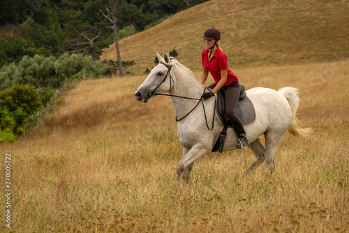 A distant shot of a beautiful happy smiling young woman dressed in a red polo shirt riding her white horse through long dried golden colour grasses, copyspace to the right