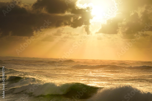The angry sea, rough waters with a setting sun behind ominous clouds, emitting rays of light through the mist onto the angry sea, crashing wave upon the rocky shore.