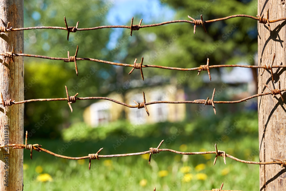 Rusty barbed wires stretched between wooden fence posts with country house on the background in summer Rusty barbed wires stretched between wooden fence posts with country house on the background in summer