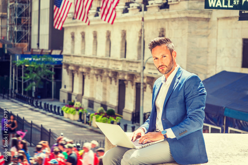 Young European Businessman with beard traveling, working in New York City, wearing blue blazer, sitting on street outside office building by Wall Street sign, working on laptop computer, thinking..