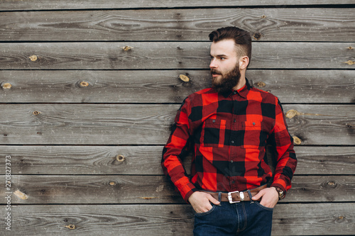 Portrait of a young stylish bearded man dressed up in jeans and a true worker black by red shirt on a wooden background.