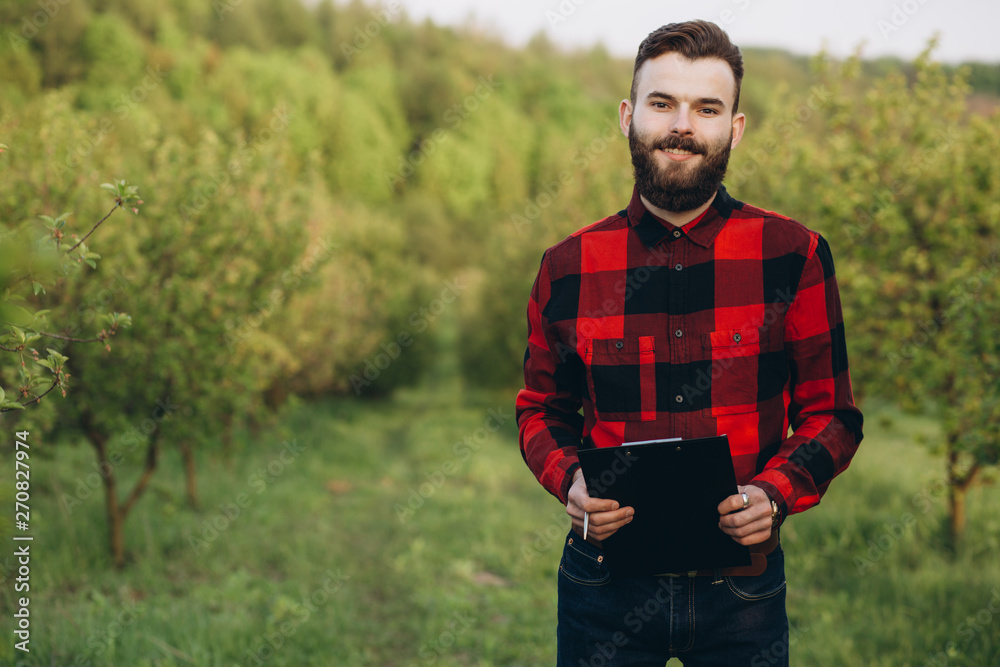 Young handsome bearded farmer with folder standing in green fruit garden in early spring