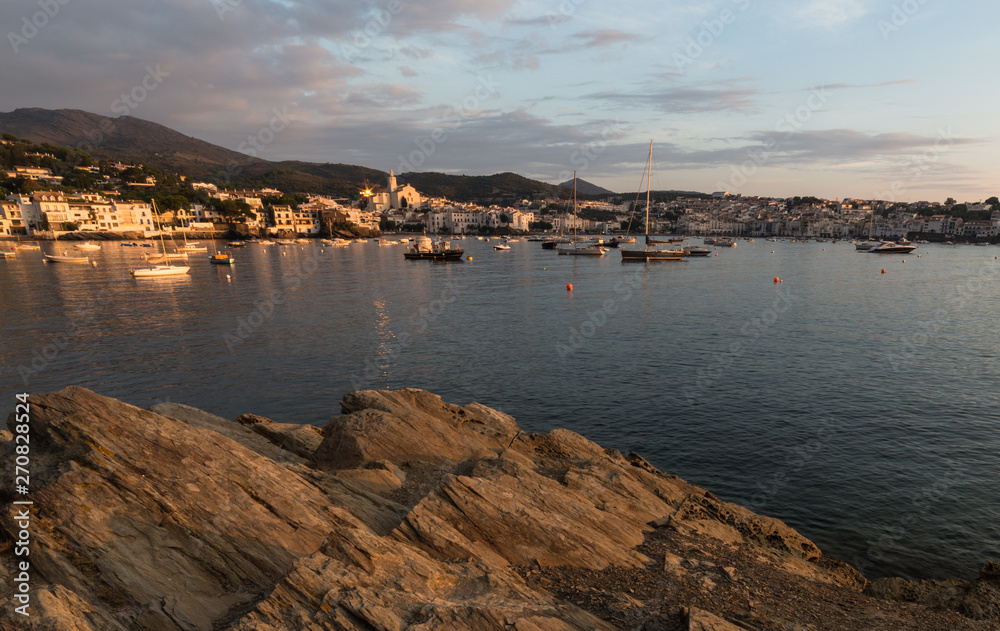 A golden atmosphere over Cadaqués. The sunrise light paints the village, boats and the rocks of the seashore.