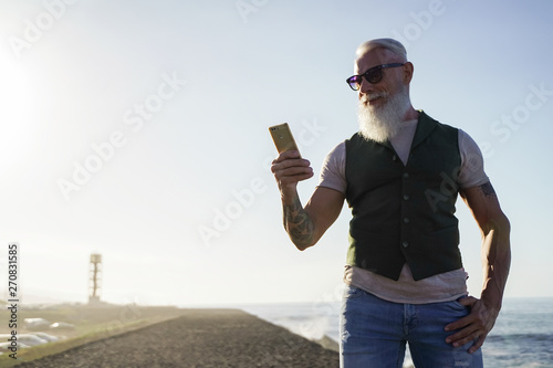 Trendy senior man using smartphone app near of the sea - Mature fashion male having fun with new trends technology - Tech and joyful elderly lifestyle concept - Focus on his face - Image