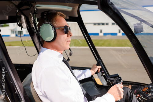 mature Pilot in sunglasses and headset sitting in helicopter cabin and holding wheel