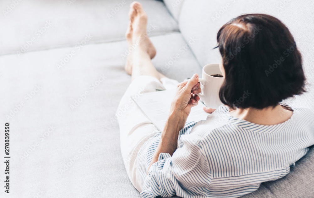 Middle-aged brunette woman on the gray sofa reading magazine with cup of coffee. Middle-aged brunette woman on the gray sofa reading magazine with cup of coffee.