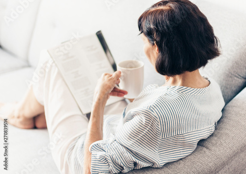 Middle-aged brunette woman on the gray sofa reading magazine with cup of coffee.