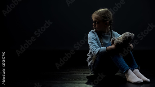Photos Little girl with teddy bear sitting alone in dark room, looking around, orphan