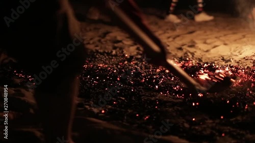 A man with a shovel shifts hot coals from a burning fire, the first man passes