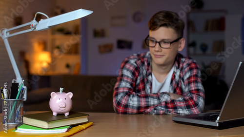 Tablou pe pânză Cheerful teenager looking into money in piggy bank, saving money for dream