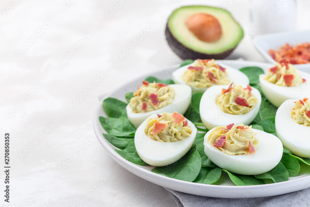 Deviled eggs stuffed with avocado, egg yolk and mayonnaise filling, garnished with bacon, on spinach leaves, horizontal copy space