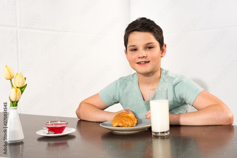 smiling boy sitting at table with croissant, syrup and glass of milk smiling boy sitting at table with croissant, syrup and glass of milk