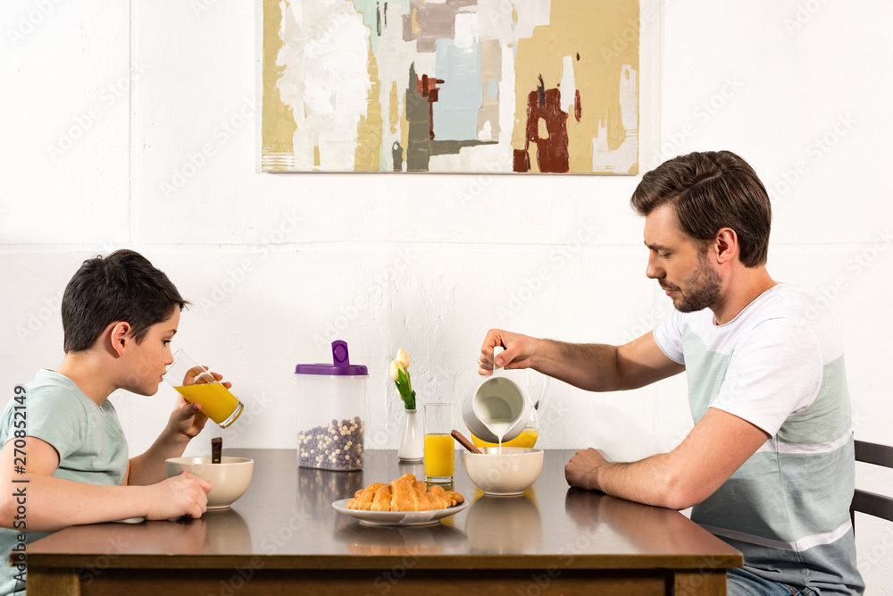 father pouring milk in bowl while sin drinking orange juice during breakfast father pouring milk in bowl while sin drinking orange juice during breakfast