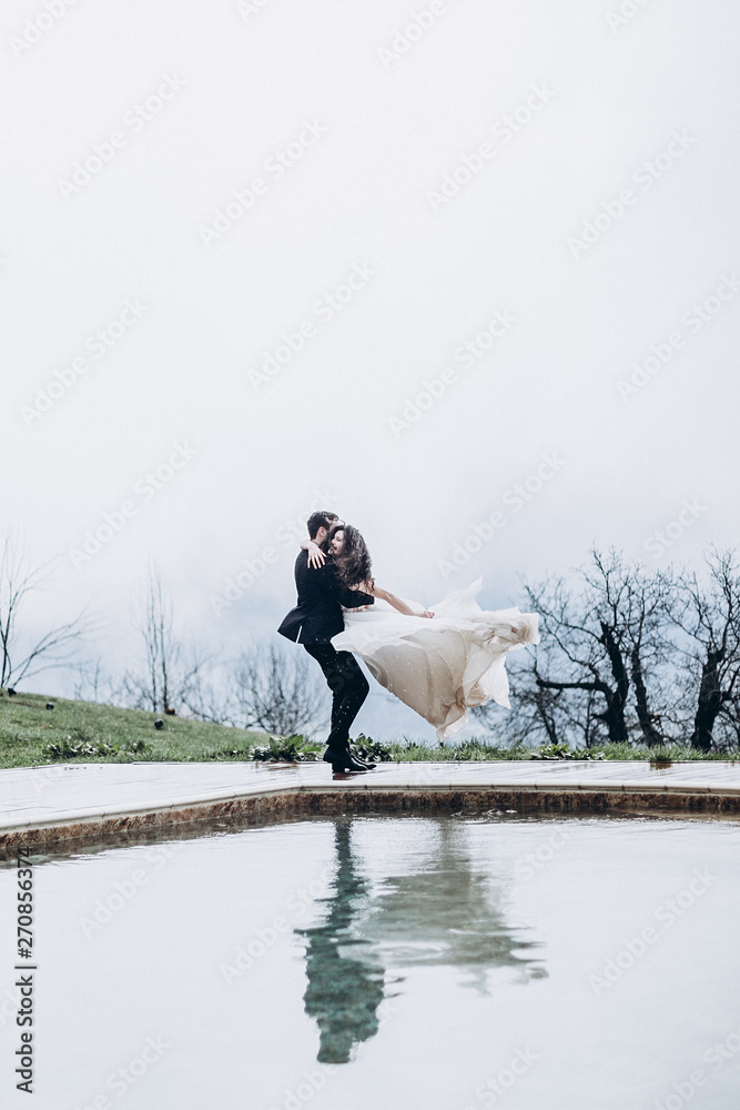 Obraz premium a stylish wedding couple photo, a man spins his bride by holding his hands near the pool in the mountains, a pair of reflections visible in the water