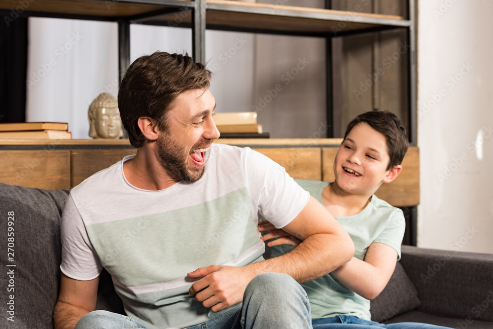 laughing son and dad sitting on sofa and having fun in living room laughing son and dad sitting on sofa and having fun in living room