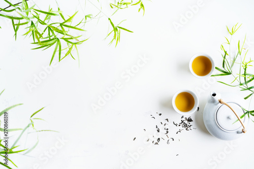 Asian tea concept, two white cups of tea and teapot surrounded with green tea dry leaves view from above, space for a text on white background.