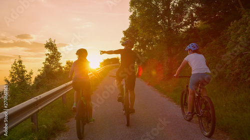 LENS FLARE: Group of cyclists rides ebikes along empty country road at sunset.