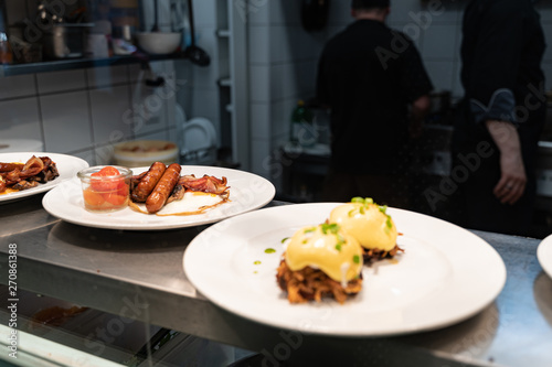 Pulled pork topped with poached egg and garnished with hollandaise sauce and breakfast sausages waiting to be served at a restaurant kitchen