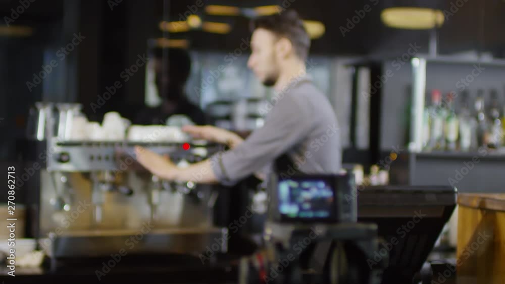 Racking focus medium shot of recording camera set on coffee shop counter, and young tattooed Caucasian barista giving demonstration of his professional coffee machine, pointing to different features