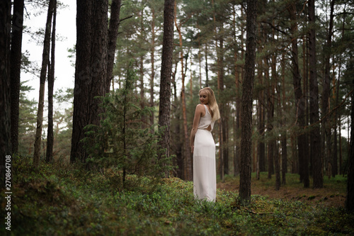 Walking Beautiful young blonde woman forest nymph in white dress in evergreen wood