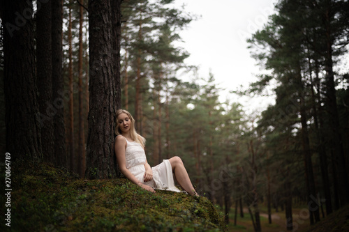 Beautiful young blonde woman sitting in forest nymph in white dress in evergreen wood © dissx