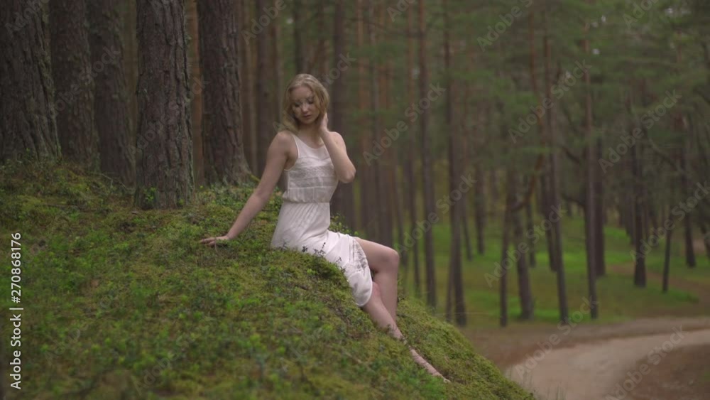 Beautiful young blonde woman sitting in forest nymph in white dress in evergreen wood Beautiful young blonde woman sitting in forest nymph in white dress in evergreen wood
