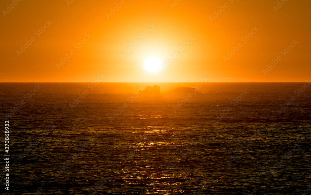 Vibrant orange sunset on the ocean with reflections on water surface and rocks in silhouette