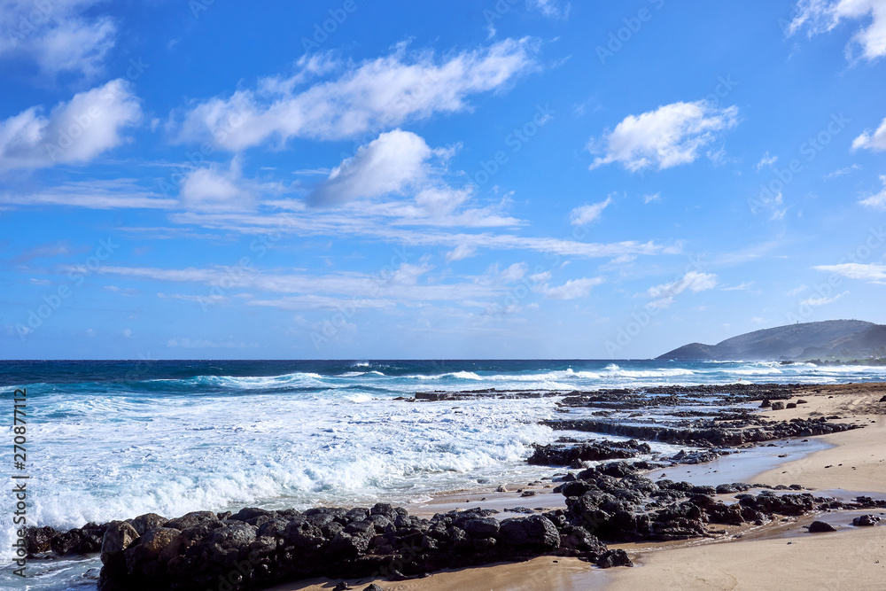 A view of Hawaiian ocean with strong waves, beautiful clouds, and rocks scattered on the beach.