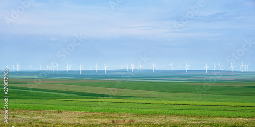 Panorama of wind turbines with layers of green wheat and rye agricultural fields, on bluish sky - space for text above and below. Concept for clean, renewable energy solutions.