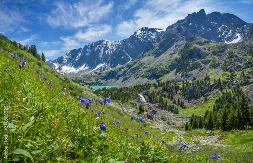 Above the picturesque mountain valley, Altai. Spring greenery, flowering slopes. A small lake under the snow-capped peaks.