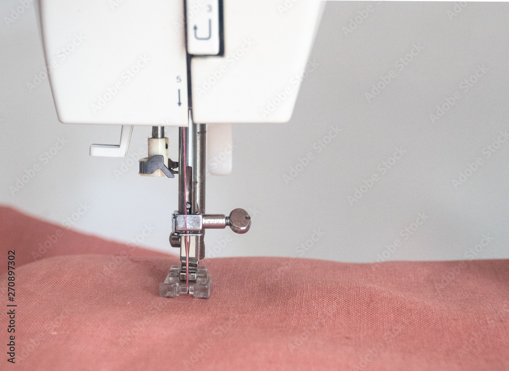 Close up of foot of sewing machine of white sewing machine while working on process with pink linen fabric on white background.