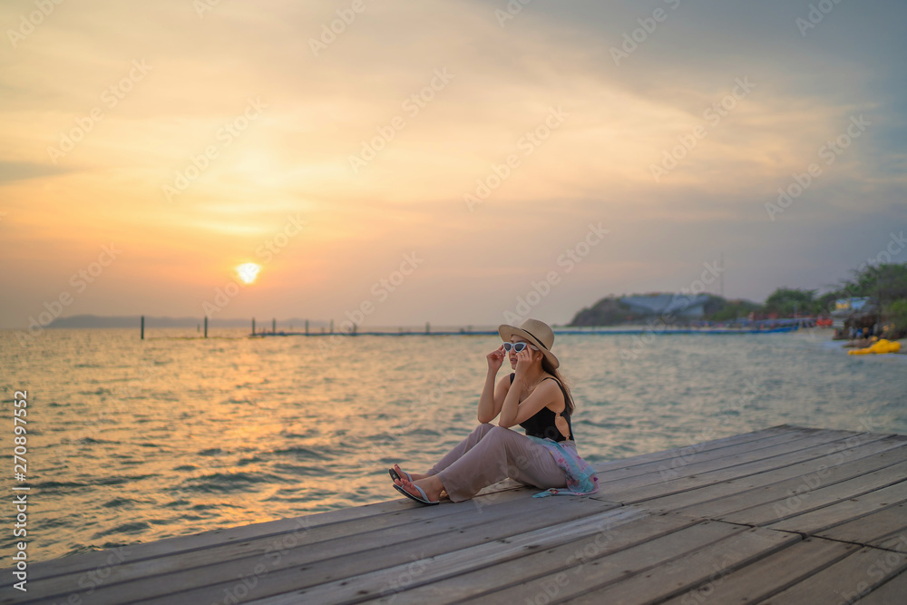 relaxing time concept with traveler girl posing smile in the sea beach in beautiful sunset time by sit on the bridge wood and wear sunglasses and hat