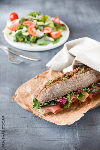 sandwich with baguette, ham, lettuce, kale on gray background, salad from kale and lettuce leaves and tomatoes on a white plate. healthy diet lunch, takeaway sandwich