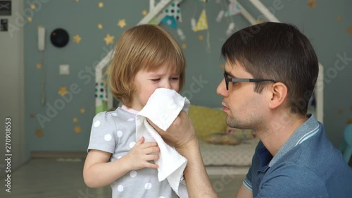 Young father with glasses wipes runny nose and tears of little cute daughter with a napkin. Baby girl cries because of illness and snot