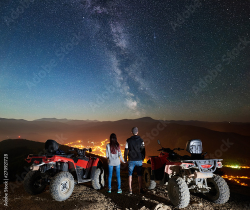 Rear view of boy and girl tourists with two atv quad motorbikes on the top of mountain, romantic couple enjoying beautiful view of night sky full of stars, Milky way, luminous town on background