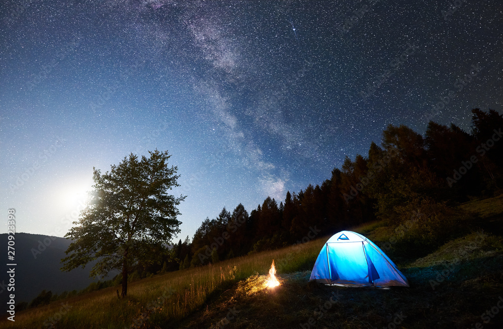 Tourist camping near forest at summer night. Illuminated tent and campfire under magical night sky full of stars and Milky way. On background big tree, beautiful starry sky, mountains and full moon