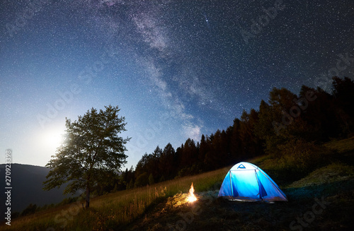 Fototapeta Naklejka Na Ścianę i Meble -  Tourist camping near forest at summer night. Illuminated tent and campfire under magical night sky full of stars and Milky way. On background big tree, beautiful starry sky, mountains and full moon