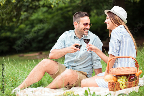 Beautiful young couple enjoying romantic picnic in a park. Relaxing on a picnic blanket and making a toast, drinking red wine. © Ivan
