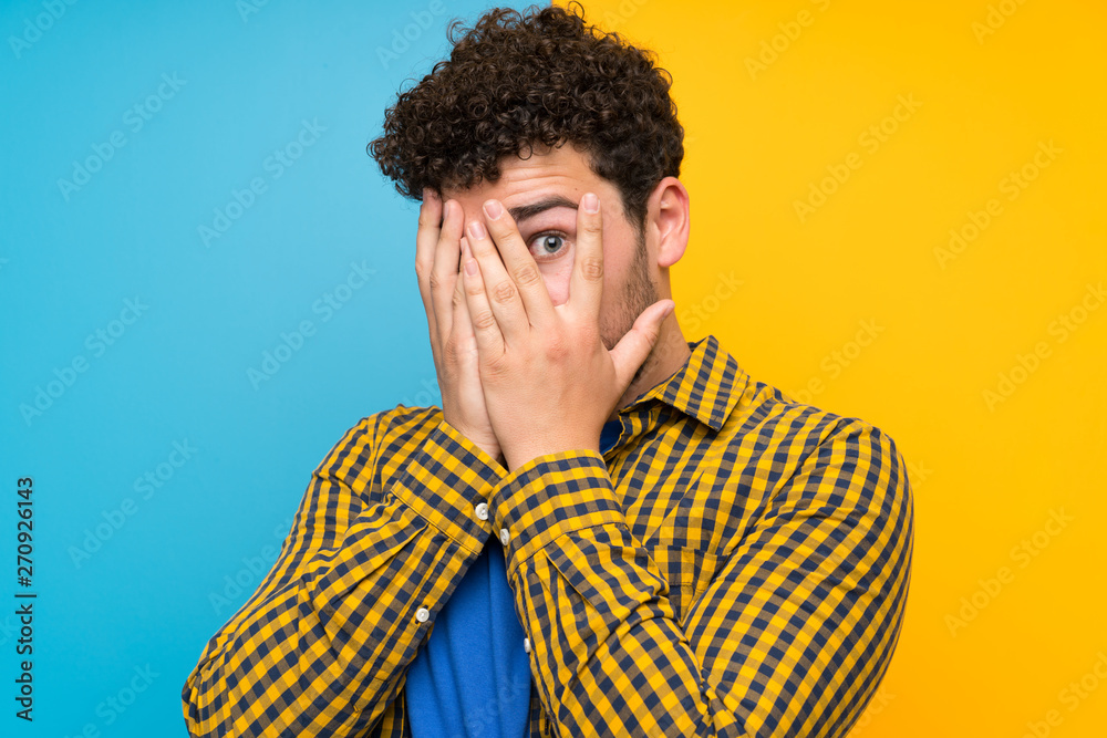 Fototapeta premium Man with curly hair over colorful wall covering eyes and looking through fingers