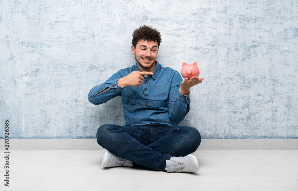 Young man sitting on the floor holding a big piggybank