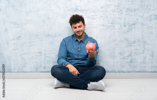 Young man sitting on the floor holding a big piggybank © luismolinero