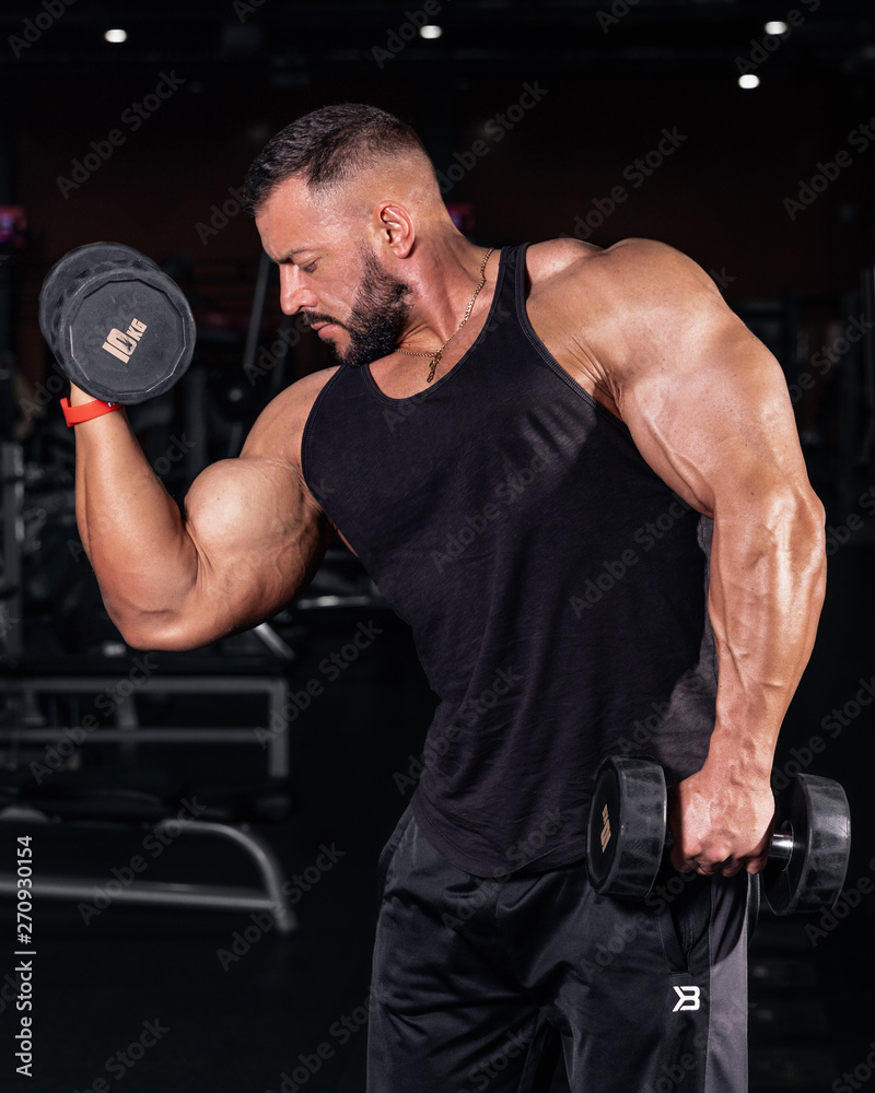 Bodybuilder in the gym. Sports photo shoot. Man's fitness. Training and exercises with dumbbells. Men's photo shoot in low key. Athletic build.