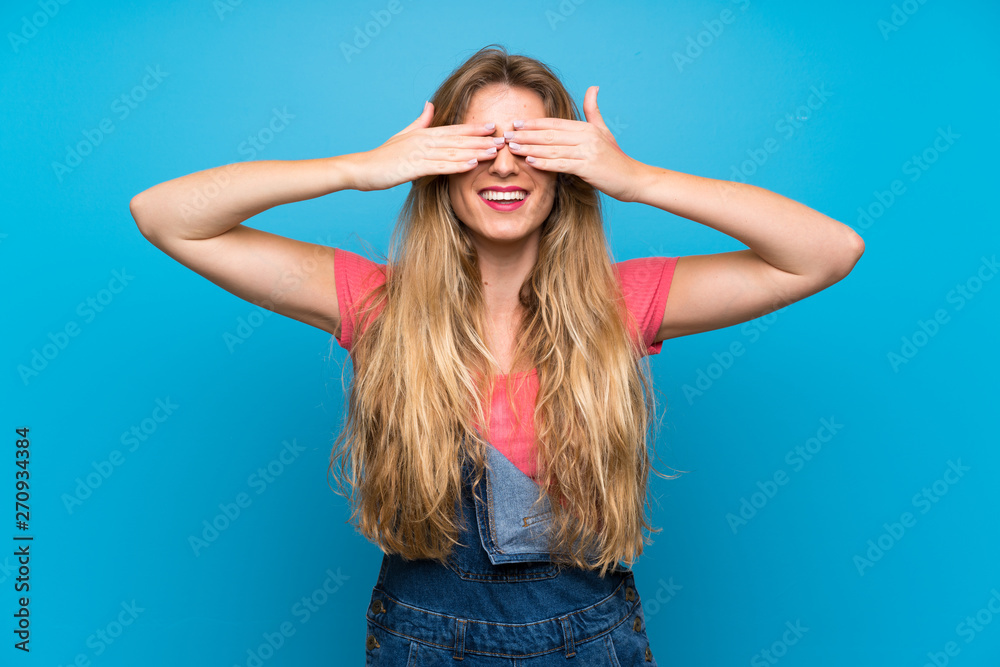 Young blonde woman with overalls over isolated blue wall covering eyes by hands