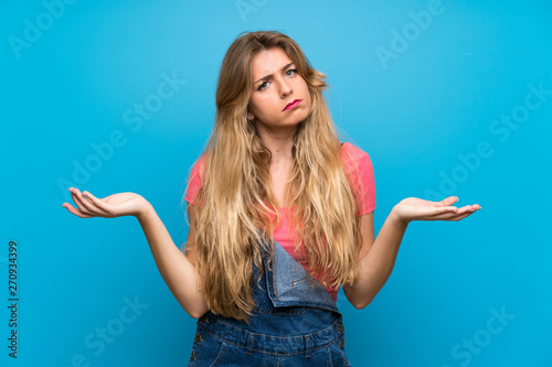 Young blonde woman with overalls over isolated blue wall unhappy for not understand something