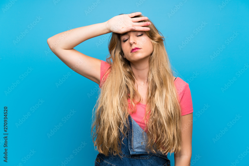 Young blonde woman with overalls over isolated blue wall with tired and sick expression Young blonde woman with overalls over isolated blue wall with tired and sick expression