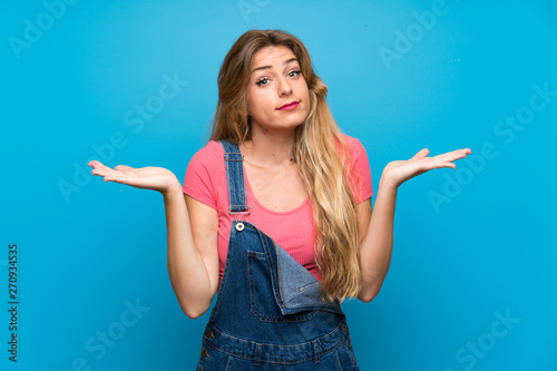 Young blonde woman with overalls over isolated blue wall making doubts gesture